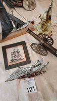 Photo showing metal sailboat with black sails, cannon model, framed ship artwork, and small shiny metal sailboat decor on a fabric surface.