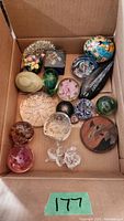 All assorted decorative items in a cardboard box shown from above, including glass paperweights, a small ceramic floral vase, a wood carved piece, and a painted trinket box.