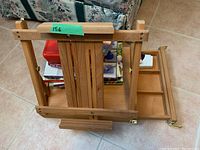 Wooden adjustable painting easel shown from the side with compartments extended and art books stacked inside and behind.