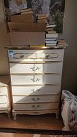 Tall cream dresser with five drawers, some books on top, floral metal handles, gold trim, vintage style