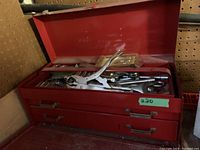 Image of an open red metal toolbox revealing various hand tools inside. Tools include pipe wrench, pliers, screwdrivers, wrenches, and miscellaneous small tools.