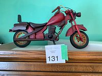 Side view of handcrafted model wooden motorcycle showing red painted wooden frame, gold-spoked wheels, and brown wooden seat.