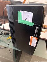 Black Oreck air purifier shown standing upright on glass table, displaying front vents and side caution stickers.