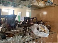 Mixed silver items including pitchers, decorative plates and a pedestal dish on a glass shelf.