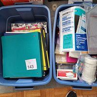 Two large plastic bins filled with assorted office supplies including colorful binders, sheet protectors, label tape, and loose papers.