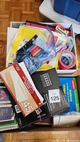 Top view showing various office supplies including notebooks, pens, markers, and packing tape in a cardboard box.