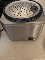 Electric rice cooker with a metal steamer insert on top shown from a front corner angle.