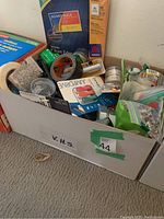 Box mainly containing Avery labels, adhesive tape rolls, and various small office supplies including pens and possibly stapler parts.
