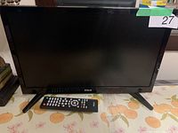 Front view of black RCA television with remote control placed in front on floral tablecloth.