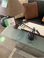 Black office table lamp on glass table with black cylindrical base and adjustable arm mechanism.
