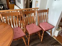 Three wooden dining chairs with spindle backs and red tufted cushions shown side by side next to a wooden dining table and railing.