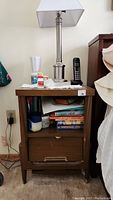Front view of one brown bedside table with items on top (lamp, phone, and bottles) and contents in shelf area.