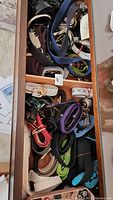 Top view of a drawer full of assorted belts, showing different colors, materials, and buckle types.