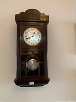Front view of the wooden wall clock with carved detail and visible clock face and pendulum.