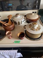 Photo showing 3 ceramic pots with lids, wooden salad bowl with wooden utensils, and two small pottery pieces on a stovetop.