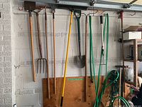 Photo showing various long-handled garden tools hanging on wall including shovels, rakes, pitchfork, hoe, and coiled green garden hose.