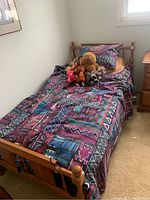 Single solid wood kids bed made of light brown wood with turned posts at the headboard and footboard, dressed with colorful bedding and several stuffed animals on top.