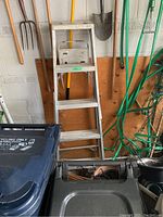 Aluminum step ladder placed against a white wall with gardening tools like pitchfork, shovel, and garden hose coiled and hanging nearby.