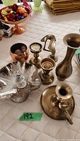 Overview of metal decorative items arranged on a table including candle holders, a vase, a small container, a figurine, and a ruffled-edge metallic plate.