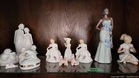 Porcelain figurines on a wooden shelf, including two ladies in detailed dresses, lady in a hat playing flute, two pink poodles, minimalist couple, and white bird group.