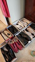 Photo of seven pairs of men's shoes including black leather, cream, tan, and red lace-up styles, all in shoe boxes, arranged on carpet.