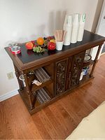 Front angled view of vintage wooden drink cart with black top holding cups and fake fruit, showing turned wooden details and carved front cabinet doors.