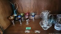 Full shelf view of six jeweled wine goblets in multiple colors and blue glass decanter