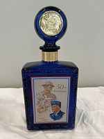 Front view of cobalt blue commemorative bottle with printed 50th anniversary American Legion label and blue stopper with gold medallion.