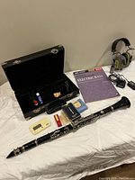 Clarinet with case open showing disassembled instrument and accessories, plus clarinet mouthpiece and reed boxes, electric bass instruction book, metronome, Ryobi headphones battery and charger.