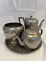 Four pewter pieces consisting of teapot, coffee pot, bucket, all placed on a round tray.