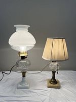 Full view of both lamps lit, showing white glass lamp with glass shade on left and clear glass lamp with fabric shade on right.