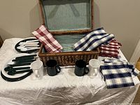 Picnic basket open displaying plastic tableware, cloth napkins, and cups set out and arranged on a white tablecloth.
