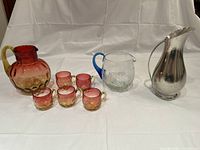 Full lot view showing three pitchers: rose glass pitcher with five matching rose cups, clear glass pitcher with blue handle, and pewter pitcher with dent.