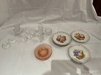 Overview of six fruit plates, four rose glass plates, and multiple clear cordial glasses and goblets on a fabric background