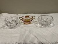 Three small bowls displayed on a white cloth: clear geometric crystal bowl, rose glass pedestal bowl with gold accents, and clear glass bowl with silver etched design.