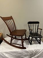 Two small rocking chairs placed together on a white sheet against a wall; one is wooden with a curved backrest and the other is black with gold accents and armrests.