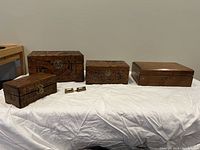 Four wooden boxes displayed side by side on a white cloth surface, three carved boxes and one plain box with two small keys visible on the cloth.