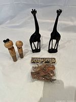 Photo showing two small wooden dolls, two tall black wooden giraffes, and a sealed set of small carved wooden animal figurines with a tag.