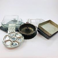 Various baking pans and kitchen tools arranged together: clear glass loaf pans with lids, metal loaf pan, Bundt pan, divided serving platter, metal BBQ chicken rack, and grater visible