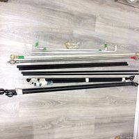 Multiple curtain rods in black, white, and medium grey with attached brackets and decorative finials, laid out horizontally on a wood grain floor.