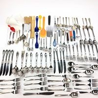 Top-down view of assorted cutlery and kitchen utensils arranged on a white surface, showing quantity and variety.