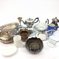 Full view of assorted silver and metal household items including bowls, teapots, jugs, platters, trays, glass containers, and ceramic pieces on white background.