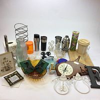 Full view of household lot items arranged on white surface including barware, decor, clock, and accessories