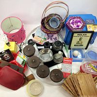 Overview photo of lot on a table including pink flamingo lamp, woven baskets, black Coors speaker, black canisters, glass bottle, red case, blue stool, and wooden craft planks.