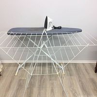 White metal drying rack with electric iron placed on top of the flat ironing board showing the setup and condition of the drying rack and iron.