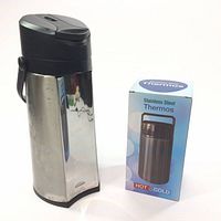 Large stainless steel thermos with black pump top and handle showing slight oxidation, beside a boxed smaller thermos