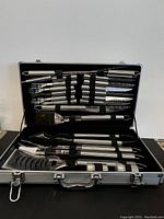 Full view of BBQ cutlery set inside aluminum carrying case showing knives, forks, tongs, brush, skewers held in black elastic loops.