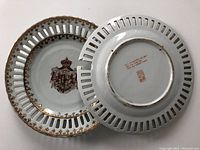 Top view showing front and back of two white porcelain pierced dishes with gold trim and central coat of arms decoration. Backside shows maker's mark.
