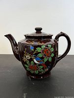Front view of vintage Asian ceramic tea pot with floral and bird painting on dark brown background.