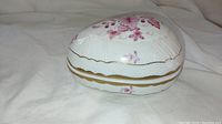 Closed view of the porcelain egg-shaped jewelry box showing pink floral and gold design on white base with subtle embossed pattern.